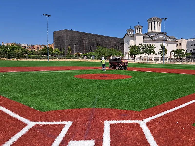 Campo de béisbol con césped artificial en proceso de instalación, con operario trabajando en el mantenimiento.