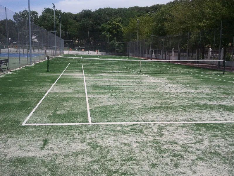 Pista de tenis de césped artificial en proceso de rehabilitación, con líneas blancas marcadas y red central.